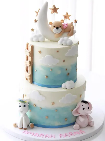A dreamy cake for twin girls' 1st birthday. This two-tiered design features a teddy bear sleeping on the moon, a unicorn, and a little pink elephant under a starry sky.