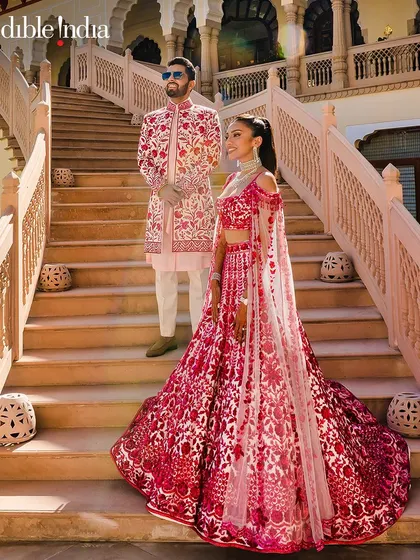 A full-length shot of the couple in their stunning red and pink outfits on the grand staircase of Rambagh Palace.