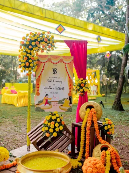 A personalized welcome to the Haldi ceremony. This setup features a custom welcome sign, colorful floral arrangements, and traditional elements like earthen pots and marigold strings.