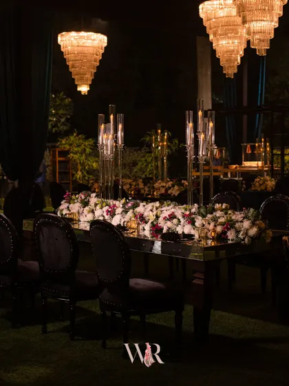 A close-up of the mirrored dining table, showing the lush floral runner composed of white, pink, and deep red blooms. The tall candle holders add height and a romantic flicker of light.