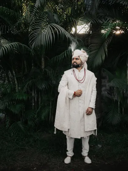 A full-length portrait of the groom standing against a dark, leafy background, looking regal.