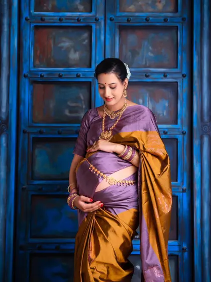 A beautiful solo portrait showcasing the elegance of a traditional Odia saree. The mom-to-be cradles her bump, standing against a vibrant blue door that makes the colors pop.