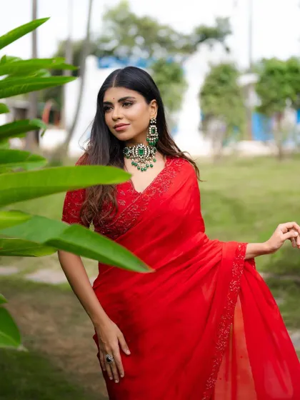 A three-quarter shot that captures the full effect of the red saree against the green foliage. The composition is clean and visually appealing.