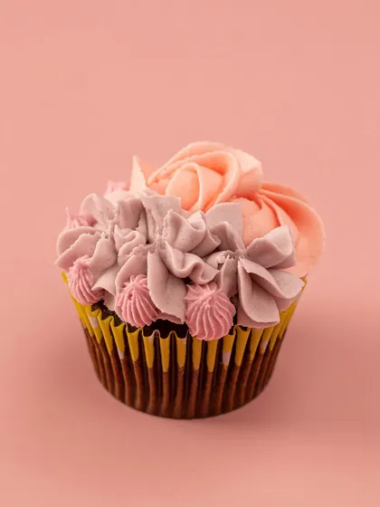 A single, beautifully crafted floral cupcake. This close-up highlights the detailed piping work, with multiple shades of pink creating a realistic, multi-petaled flower on a Belgian chocolate base.