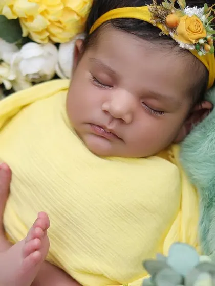 A close-up portrait focusing on the baby's tiny feet, peeking out from a sunny yellow swaddle. The floral headband and surrounding flowers complete this cheerful setup.