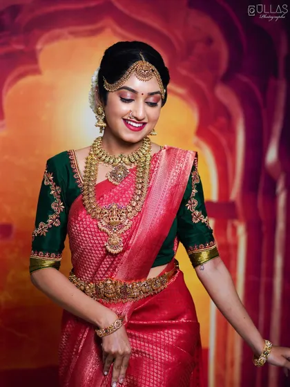 A full shot of the bride in her red and green silk saree, adorned with a complete temple jewellery set including a choker, haaram, and waist belt.