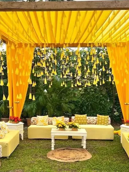 An outdoor lounge setup for a Haldi or Mehendi function. The yellow drapes and hanging floral tassels create a cheerful and relaxed canopy for guests to sit under.