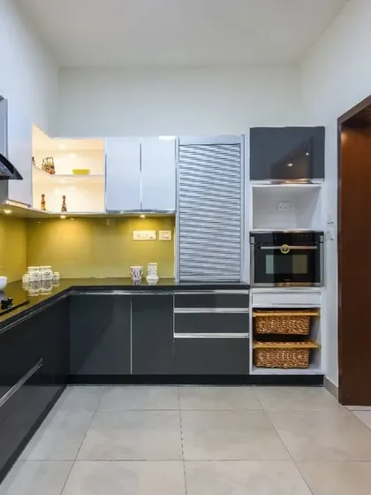 A U-shaped kitchen with glossy dark grey cabinets and a mustard-yellow backsplash. The design includes a tall unit with a rolling shutter and wicker baskets for vegetable storage.