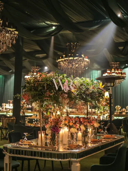 A stunning mirrored table reflects the grand floral centerpiece and the warm glow of candles. Set under a ceiling of green drapes and chandeliers, this tablescape was a focal point of Nikita and Pushkar's elegant engagement dinner.