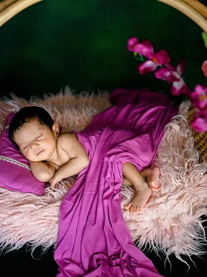 A newborn sleeping peacefully in a hanging basket, draped in a rich purple cloth. The dark background makes the colors stand out.