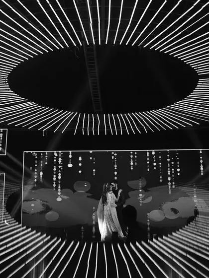 A wide shot of the couple on a massive, circular stage with stunning lighting design. This image highlights the grand production value and cinematic quality of modern Sangeet nights.
