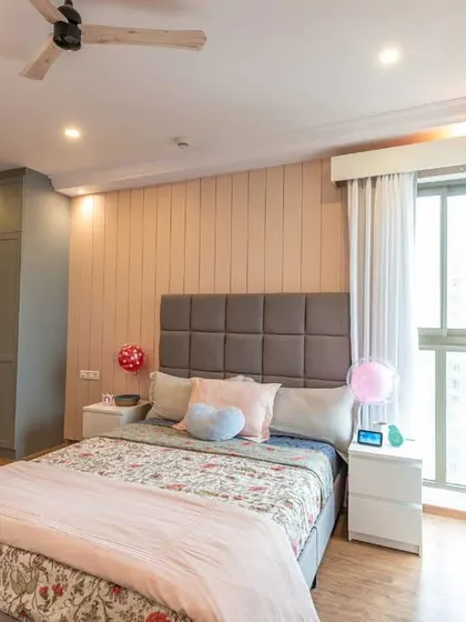 A cozy bedroom for a girl, featuring a cushioned headboard, soft pink paneling, and a white study unit with display shelves for personal treasures.