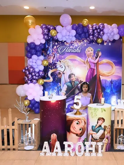 A magical Rapunzel-themed birthday party for a 5-year-old. The decor features a large backdrop with characters from Tangled, a purple and gold balloon garland, and themed props.