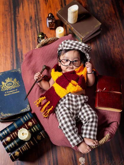 A little wizard in the making. This baby is all ready for Hogwarts, dressed in a plaid outfit with a Gryffindor scarf and round glasses, holding a wand in a magical, book-filled setting.