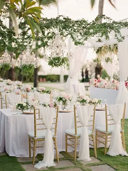 An elegant outdoor wedding reception setup. The tables are beautifully set with floral centerpieces, and the chairs are decorated with flowing white drapes.