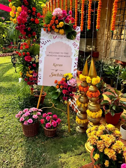 A welcome sign for Karan and Suchita's Mehendi, surrounded by colorful flowers and traditional temple-style brass decor.