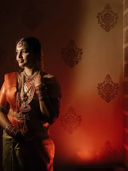 A dramatic bridal portrait using strong side lighting. The bride stands against a textured wall, her traditional South Indian attire glowing in the warm light.