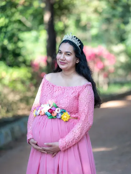 A classic princess-style portrait. This mom-to-be wears a tiara and a floral belt with her pink gown, looking serene in a beautiful park setting.