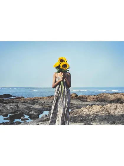 A whimsical shot for the 'Sacred Wrap' campaign for Adiv x Kavita Parmar. The styling is playful, with the model's face hidden by sunflowers, set against the raw, natural beauty of a rocky shoreline.