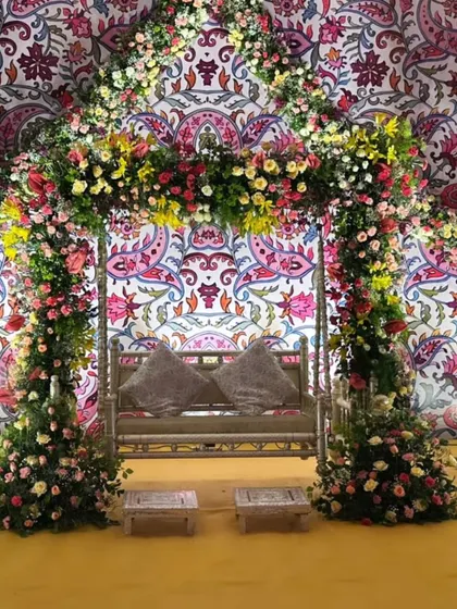 A beautiful Mehendi seating area with a floral-draped swing set against a vibrant, patterned backdrop, flanked by elegant white peacock statues.