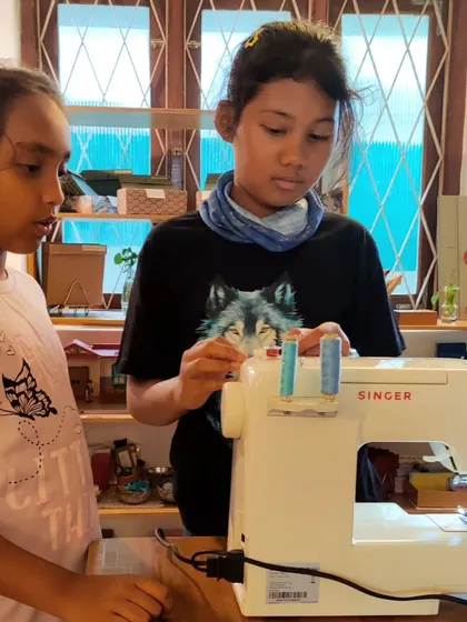 An older child mentors a younger one, showing her how to thread the sewing machine. This is a beautiful example of the natural teaching and learning that happens in our mixed-age environment.