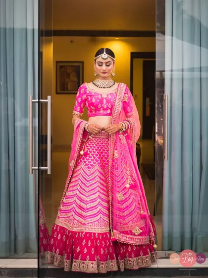 A full-length portrait of the bride, Mohi, looking stunning in a vibrant pink lehenga as she stands in a doorway.