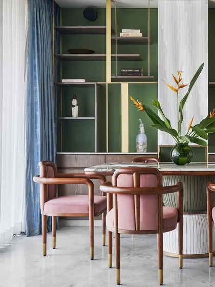 The dining area of the "Whimsical House," featuring a custom shelving unit with brass frames that houses the client's cherished art pieces. The 'Svelte' chairs in mauve add a soft, luxurious touch.