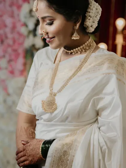 A close-up of the bride's portrait, focusing on her serene expression and the exquisite details of her traditional gold jewelry and white wedding saree.