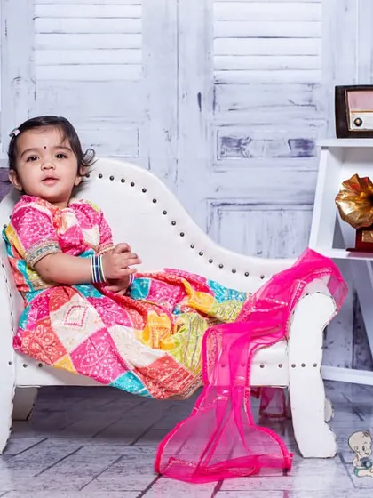 A toddler looking elegant in a colorful, traditional dress, seated on a white chaise lounge. The vintage-inspired props and setting create a classic and graceful portrait.