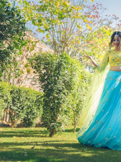 A bride enjoying a moment in a sunlit garden. Her vibrant yellow and blue lehenga for a Mehendi ceremony looks stunning against the natural greenery.