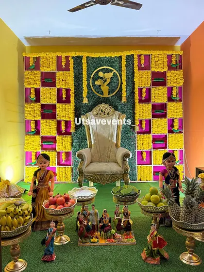 A home baby shower setup with a checkered marigold backdrop and a golden throne. The arrangement includes traditional dolls and offerings on brass stands.