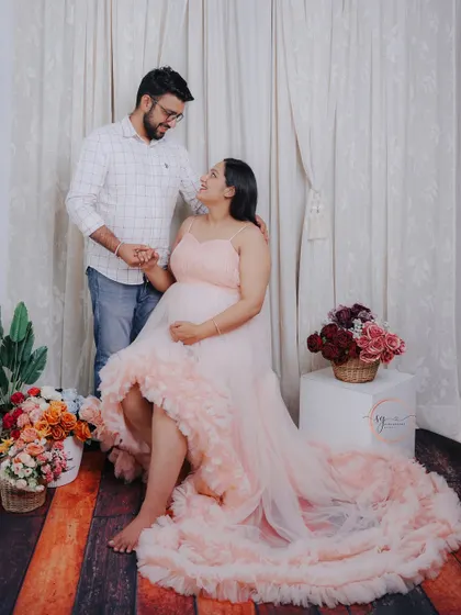 A romantic studio maternity photo with a soft, dreamy feel. The couple shares a loving gaze amidst a setup with flowing drapes and floral accents, creating a truly magical atmosphere.