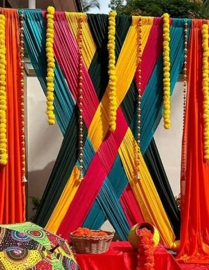A creative and modern backdrop design using a criss-cross pattern of colorful drapes in orange, yellow, teal, and black. This geometric approach to fabric draping is accented with marigold strings for a fusion look.