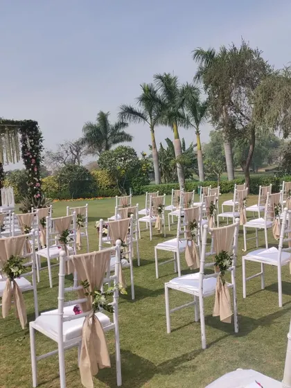 This is a guest seating arrangement for an outdoor wedding ceremony. The white tiffany chairs are elegantly decorated with simple fabric ties and small floral bunches, facing the mandap area.