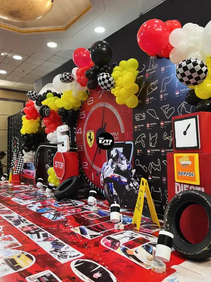 A close-up of the main stage for a Ferrari-themed birthday. The backdrop features a race track map, a giant clock, and balloon garlands in the signature red, yellow, and black colors.