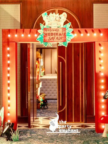 The entrance to the jungle safari party was designed to look like a park gate, with a lit-up red frame, a 'Welcome to Vedika's Safari' sign, and a friendly zebra prop to greet guests.