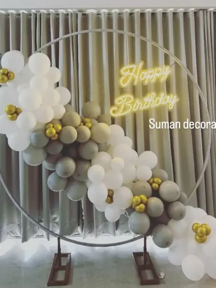 A simple and modern birthday setup with a grey, white, and gold balloon garland on a circular frame. The 'Happy Birthday' neon sign adds a warm glow.