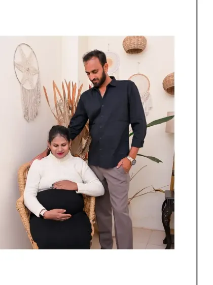 A sweet and supportive couple's portrait. The partner stands behind the seated mom-to-be, creating a sense of protection and togetherness. The natural textures of the wicker chair and wall hangings enhance the warm atmosphere.