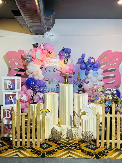 A magical unicorn-themed first birthday celebration. This setup features large butterfly wing cutouts, pastel balloon decorations, unicorn balloon pillars, and a full set of props including a picket fence and 'BABY' blocks.