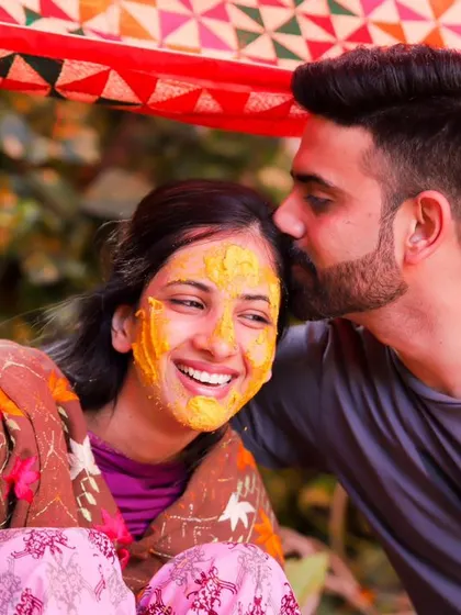 A tender and happy moment between the couple at their Haldi. His arm is around her, and he gives her a sweet kiss on the forehead as she smiles.