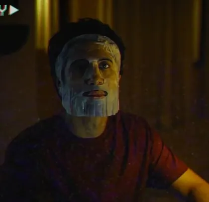 A playful scene from The Man Company film. The face mask adds a touch of humor, and the casual red t-shirt is part of the character's relaxed, at-home wardrobe.