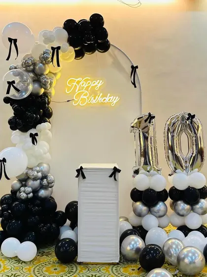 A chic and modern 10th birthday with a black and white bow theme. This setup includes a balloon arch, a neon sign, and number balloons for a stylish celebration.