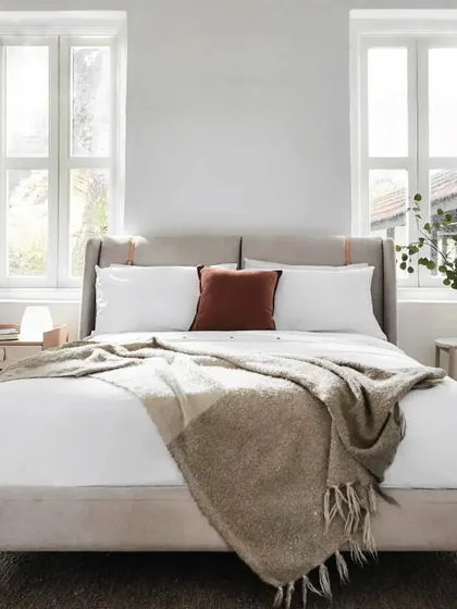 The Crème Bed, set in a bright, airy room, embodies classic Scandinavian design. The fabric-upholstered headboard with leather strap accents and tapered oak legs creates a look that is both clean and calming.