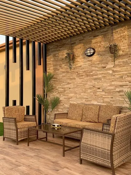 A rustic patio design with wicker furniture set against a stacked stone wall. The slatted wooden ceiling provides shade and adds to the natural, earthy feel of the space.