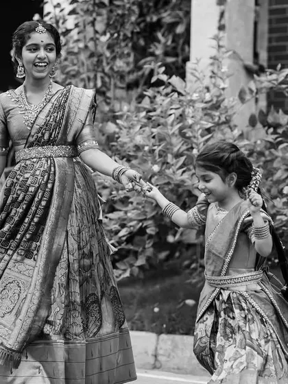 A beautiful black and white photo of an older sister or bridesmaid leading a flower girl, both dressed in traditional half-sarees.