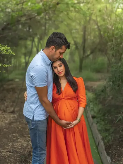 A tender outdoor portrait of a couple. The husband gently kisses his wife's head as they both cradle her belly, surrounded by the soft greenery of the woods.