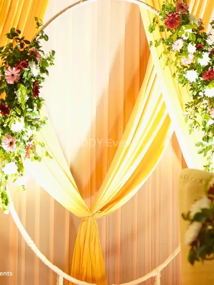 This unique stage backdrop features a large white ring adorned with floral arrangements and draped with yellow fabric. It offers a modern and artistic alternative to traditional stage designs for an engagement or Haldi ceremony.
