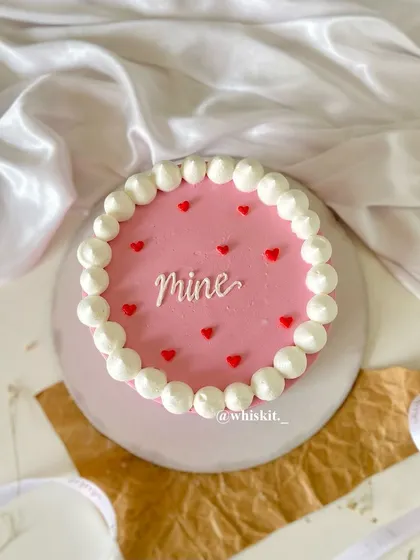 A sweet and simple pink flat cake with "Mine" written on it, surrounded by a border of piped pearls and tiny red hearts.