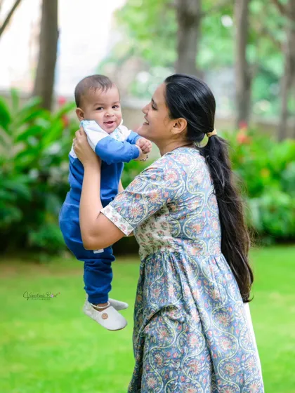 A mother lifts her smiling baby high in an outdoor setting. The joy on both their faces is infectious, making this a wonderful, candid family photograph.