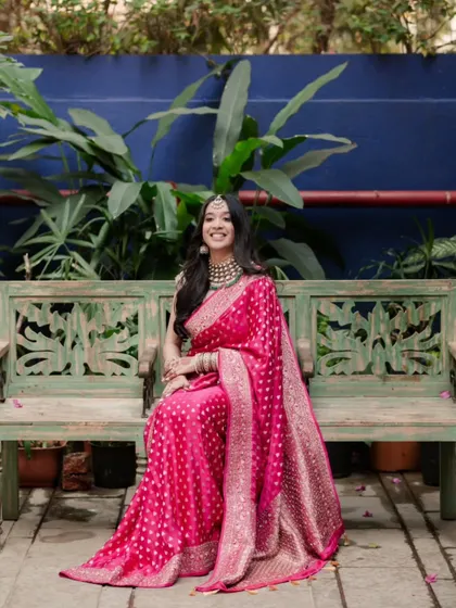 The bride, dressed in a vibrant pink saree, smiles brightly while seated on a rustic bench, a perfect blend of elegance and joy.